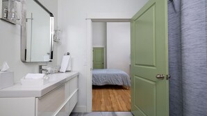 A modern bathroom with a white vanity, a large mirror, and a green door leading to a bedroom with a wooden floor and a bed with a patterned bedspread. - Lou-City Penthouse by Hollyhock Suites (Louisville)