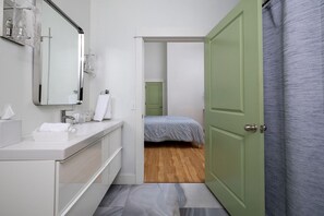 Un baño moderno con un tocador blanco, un espejo grande y una puerta verde que da a un dormitorio con suelo de madera y una cama con colcha estampada.