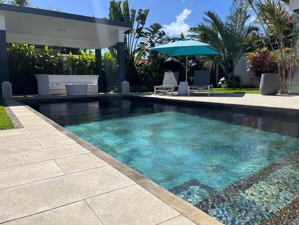 A swimming pool with a tiled deck, lounge chairs, and a covered seating area.