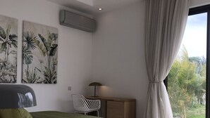 A modern bedroom with a large bed, a desk, and a chair. There is a window with curtains and a view of greenery outside.