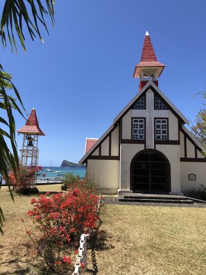 A church with a red roof and a bell tower, surrounded by a grassy area with red flowers and a clear blue sky.