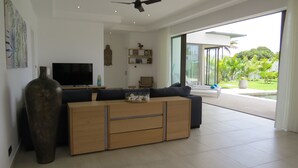 A modern living room with a large TV, a wooden cabinet, a dark sofa, and a view of a lush garden through large windows.