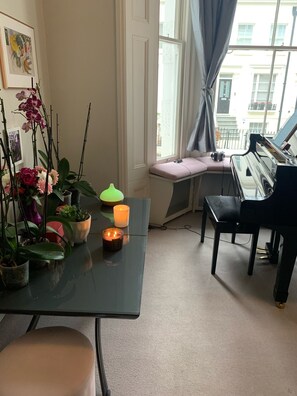 A room with a piano, a table with plants and candles, and a window with a view of buildings.