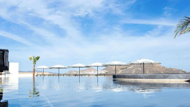 An infinity pool with umbrellas and palm trees, reflecting a mountainous landscape. - 2 BR Stunning Ocean Views Condo · Copala Quivira (Cabo San Lucas)