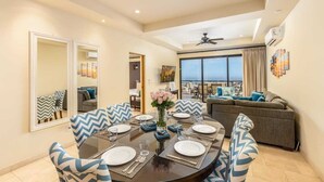 A dining area with a round table set for four, a sofa, and a view of the city. - 2 BR Stunning Ocean Views Condo · Copala Quivira (Cabo San Lucas)