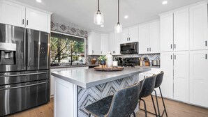 A modern kitchen with white cabinets, stainless steel appliances, a central island with bar stools, and pendant lighting. - Modern Mountain Cabin w/ Hot Tub, Game Room & Fire Pit (Big Bear)