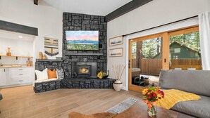 A living room with a stone fireplace, a sofa, a coffee table, and a view of the outdoors. - Modern Mountain Cabin w/ Hot Tub, Game Room & Fire Pit (Big Bear)