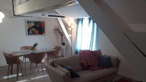 A modern living room with a beige sofa, a glass dining table with pink chairs, a painting on the wall, and a staircase leading to an upper level. - Apartment T3 ❤de ville. (La Rochelle)