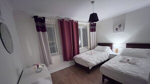 A bedroom with two beds, a bedside table with a lamp, a window with curtains, and a wall-mounted picture. - River Bay (Cardiff)