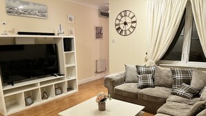 A living room with a flat-screen TV, white shelves, a grey sofa with plaid cushions, and a wall clock. - River Bay (Cardiff)