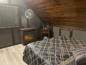 Ett sovrum med ett sluttande tak, en vedkamin, en säng med ett mönstrat överkast, ett nattduksbord och en väggmonterad fläkt.