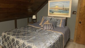Een slaapkamer met een schuine plafond, een bed met een bedovertrek met een patroon, een nachtkastje met een lamp en een lijst met een schilderij van een kustlandschap.