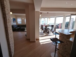 Ampla sala de estar com parede de destaque em pedra, sofá escuro e área de jantar com mesa e cadeiras.