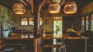 A rustic kitchen with wooden countertops, hanging wicker lights, and a view of the outdoors through large windows.