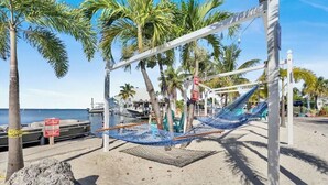 Een hangmat tussen palmbomen op een zandstrand, met een steiger op de achtergrond.
