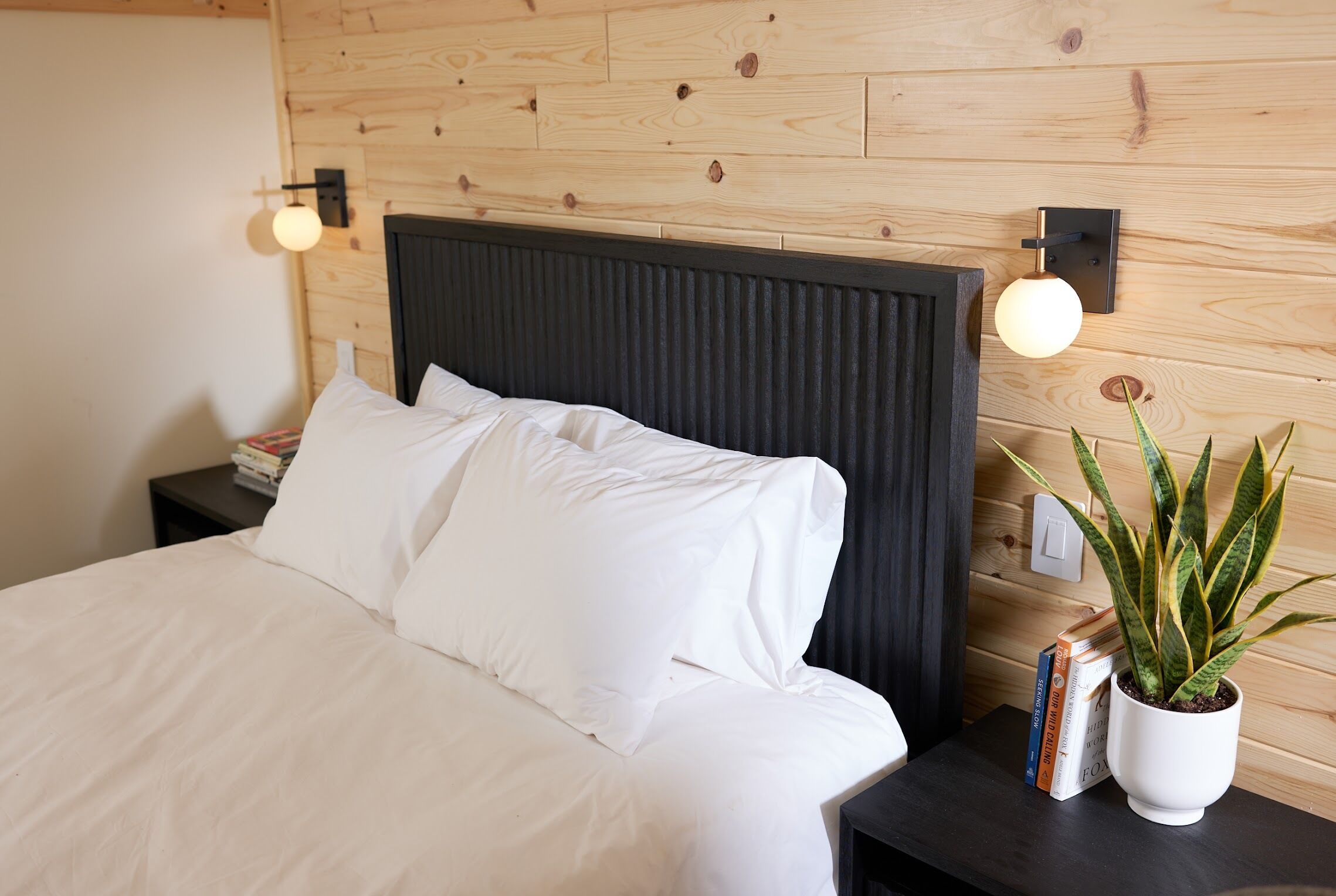A neatly made bed with white linens, a dark headboard, and a bedside table with a potted plant and books.