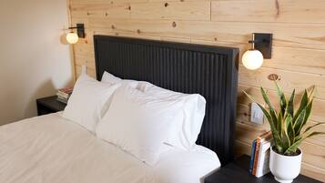 A neatly made bed with white linens, a dark headboard, and a bedside table with a potted plant and books.
