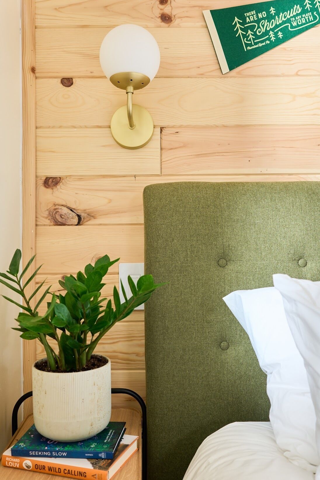 A neatly arranged room with a green upholstered headboard, a bedside lamp, a potted plant, and a few books on a small table.
