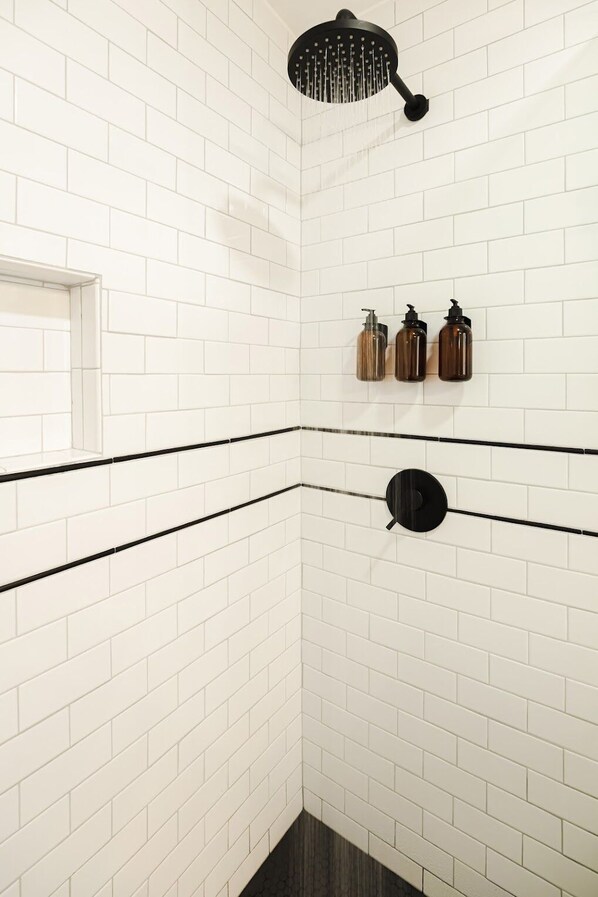 A corner bathroom with white subway tiles, black trim, a showerhead, and three bottles of soap on the wall. - The Rex Hotel (Greentown)