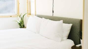 A neatly made bed with white linens in a room with a window, a plant, and a lamp.