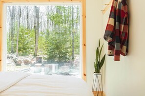A bedroom with a large window overlooking a forest, a bed, a plant, and a hanging coat rack. - The Rex Hotel (Greentown)