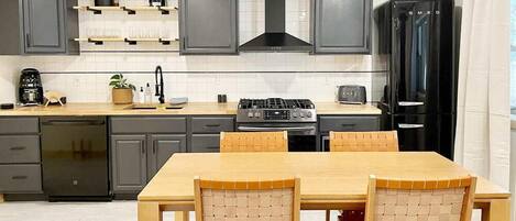 A modern kitchen with dark grey cabinets, a black range hood, and a black refrigerator. There is a wooden dining table with four chairs.