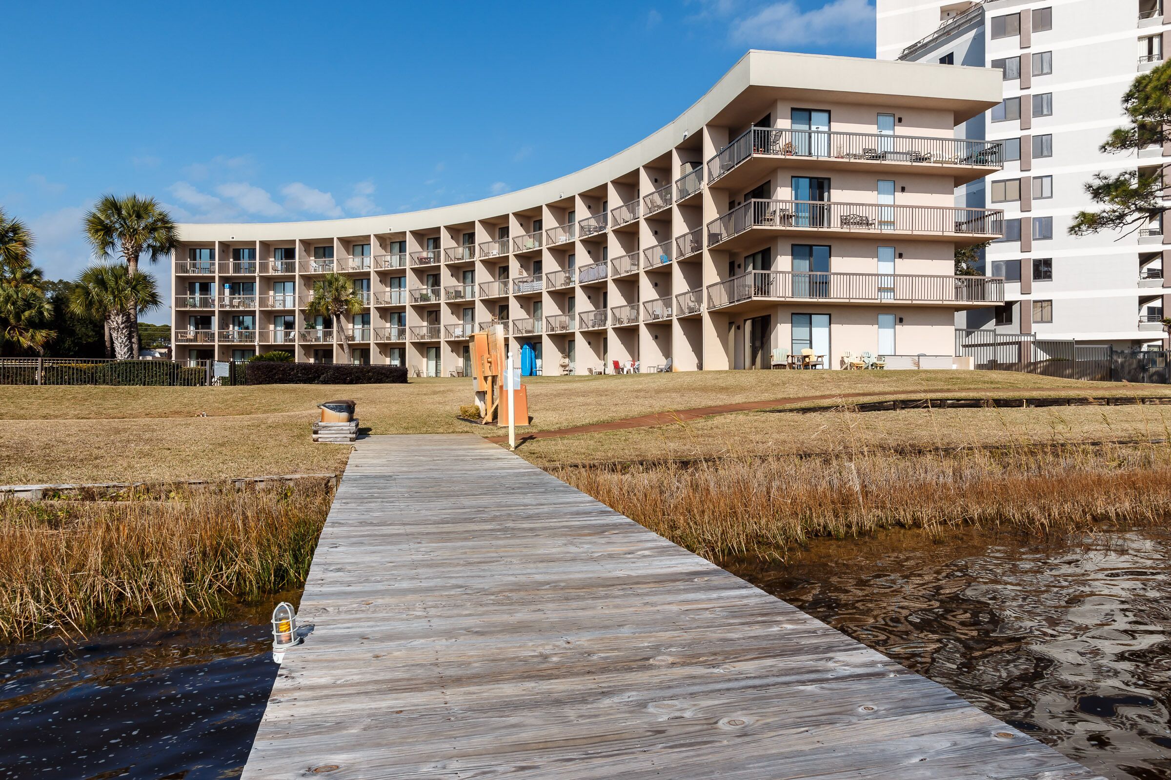 Un hôtel moderne à la façade incurvée, doté de nombreux balcons et d’une promenade en bois menant à un espace verdoyant.