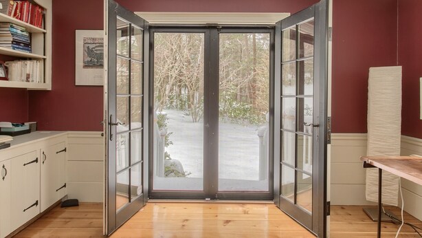 A room with a sliding glass door leading to a snowy outdoor area, a white cabinet with books, and a wooden floor. - New Luxury Listing: 5,000 sq/ft Secluded House (Weston)