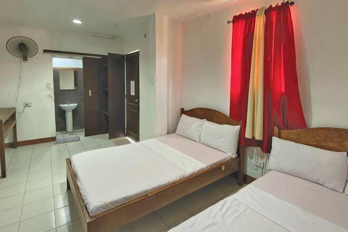 Een hotelkamer met twee bedden, een bureau, een stoel, een ventilator en een badkamer die door een geopende deur zichtbaar is.