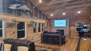 A cozy, rustic interior with a wooden ceiling, a large TV screen displaying 'Southwest.com', a bar area, and a seating area with leather chairs. - 3 Bedroom Loft With Game Room ~ In The Heart Of Downtown Copper Harbor (Copper Harbor)