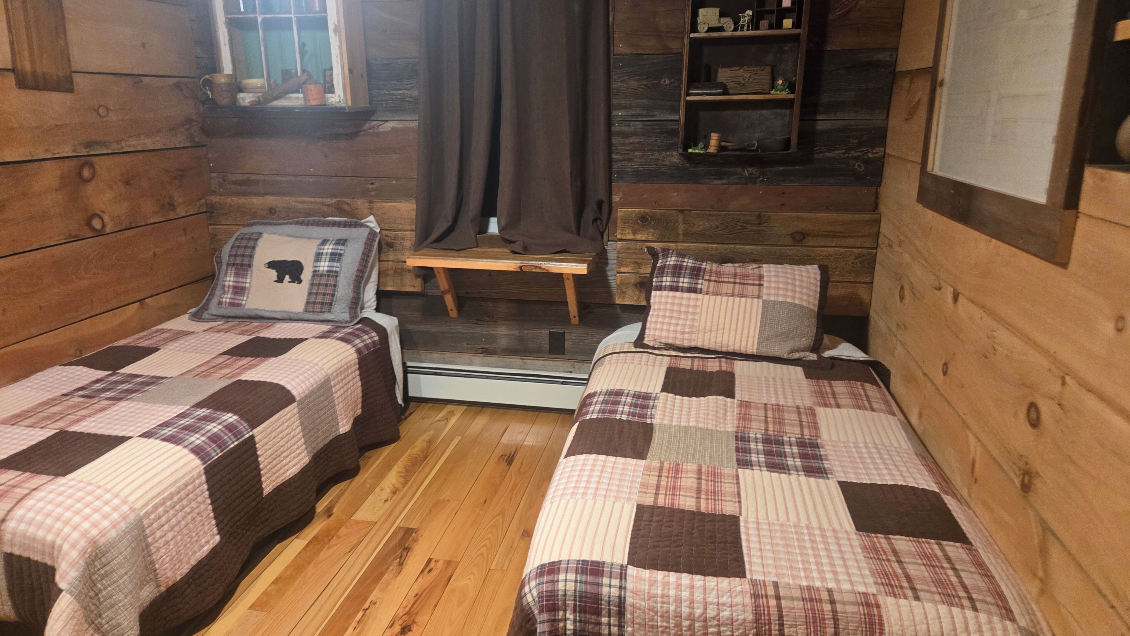 Una cabaña con dos camas, una mesita, un estante con objetos y una ventana con cortinas.