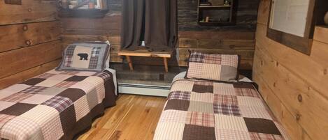 Una cabaña con dos camas, una mesita, un estante con objetos y una ventana con cortinas.