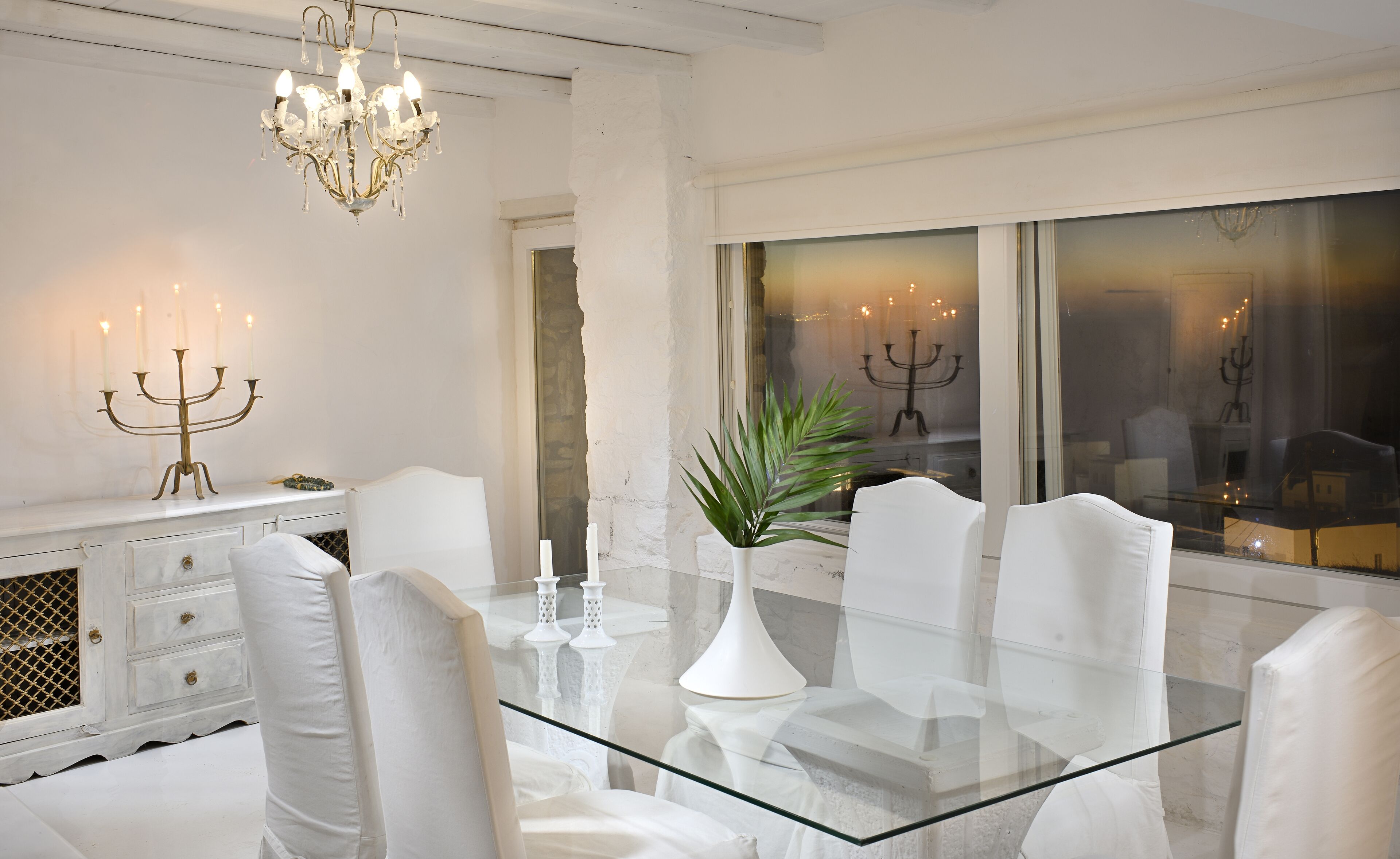 Een moderne eethoek met een glazen tafel, witte stoelen en een kroonluchter.