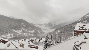 A snow-covered mountain village with traditional chalet-style buildings, a valley, and a lake in the background.