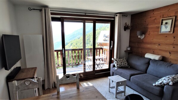 A cozy living room with a large sliding glass door leading to a balcony, a grey sofa, a small wooden table, and a mounted TV.