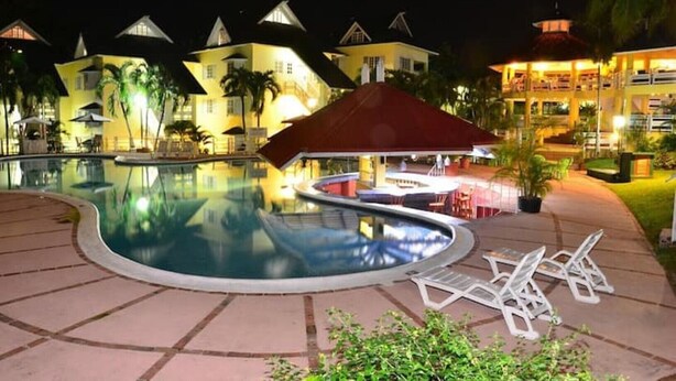 A nighttime view of a resort with a swimming pool, lounge chairs, and a covered area with tables and umbrellas. - 1 Bedroom Penthouse Suite 63, Mystic Ridge Resort (Ocho Rios)
