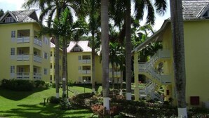 A yellow multi-story hotel with balconies, palm trees, and a well-maintained garden. - 1 Bedroom Penthouse Suite 63, Mystic Ridge Resort (Ocho Rios)