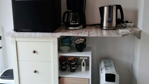 Una kitchenette compacta con refrigerador, cafetera, hervidor y un ventilador pequeño.
