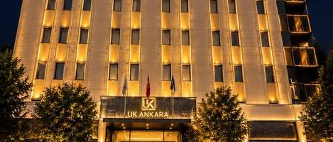Un hotel de varios pisos con una entrada destacada, un estacionamiento con varios automóviles y una fachada bien iluminada.