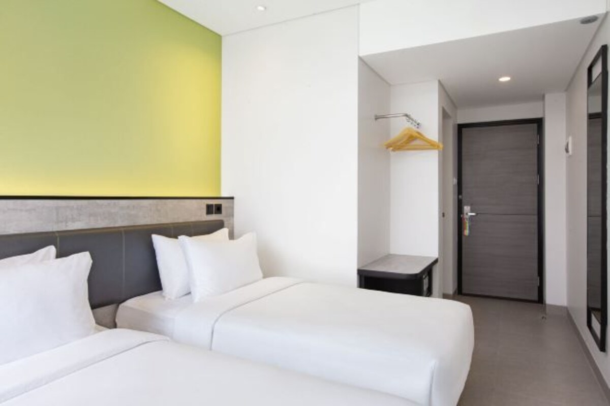 A hotel room with two beds, a small table, a hanging coat rack, and a door.