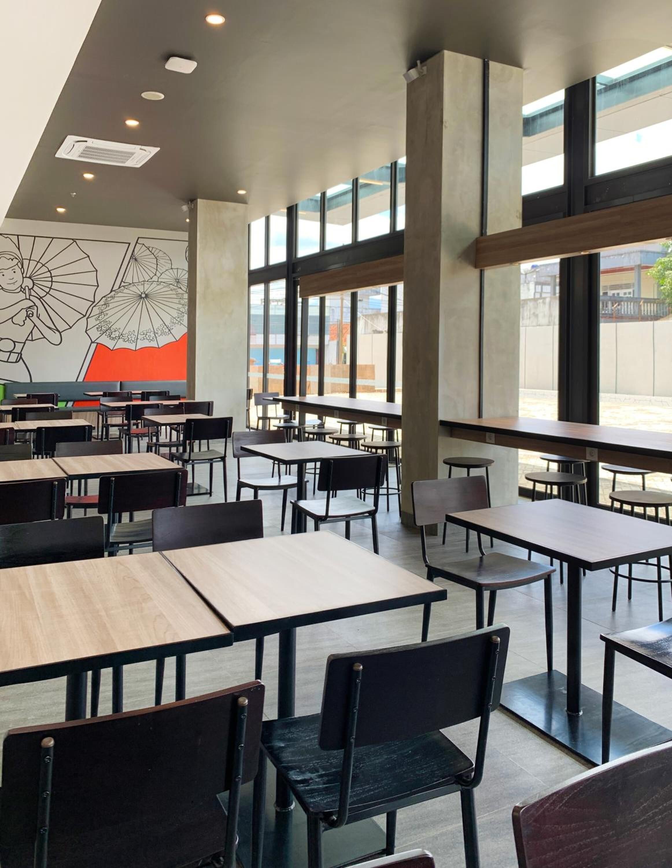 A modern dining area with high ceilings, large windows, and a mural of umbrellas. Tables and chairs are arranged for dining.