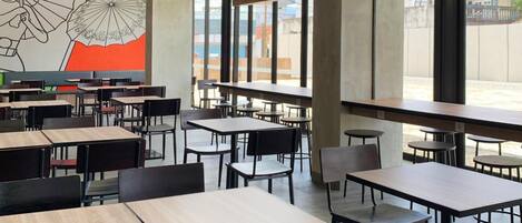 A modern dining area with high ceilings, large windows, and a mural of umbrellas. Tables and chairs are arranged for dining.