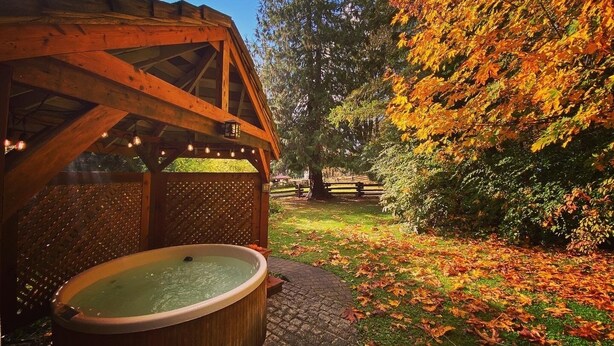 Un quiosco de madera con jacuzzi, rodeado de un jardín con hojas otoñales y una cerca de madera.