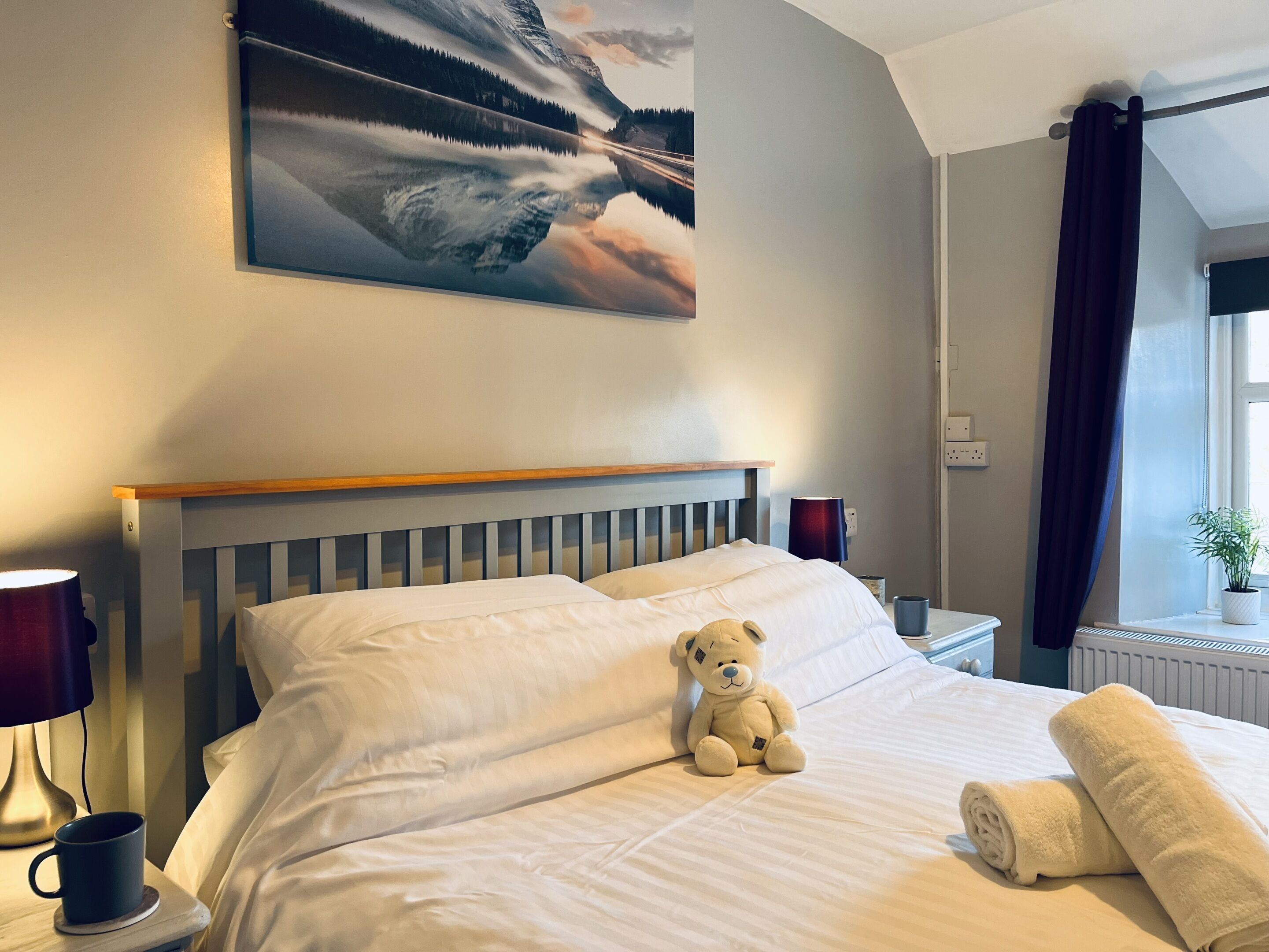 Quarto de hotel com cama, um ursinho de pelúcia, toalha enrolada, abajur e uma pintura de paisagem montanhosa.
