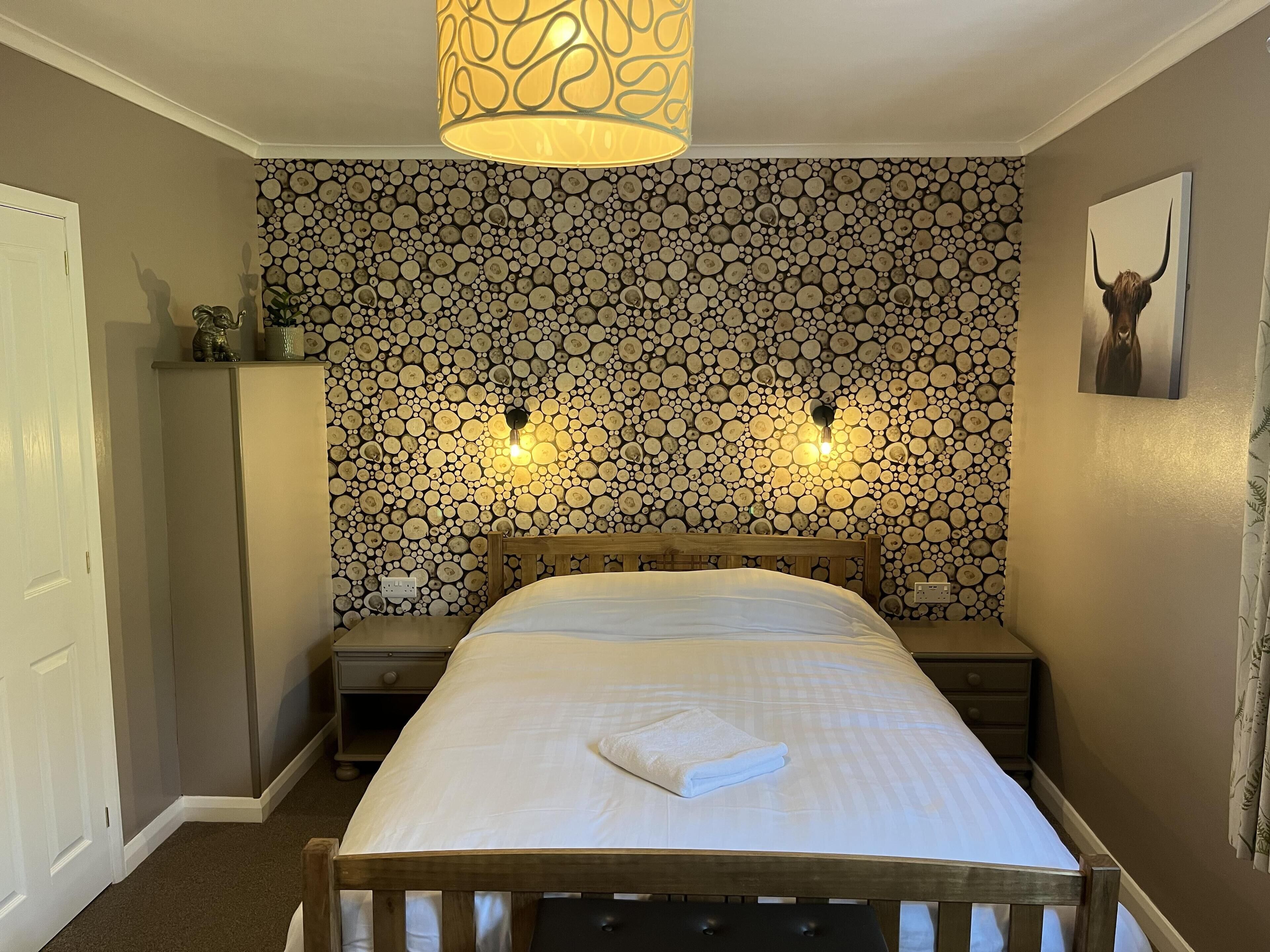 Une chambre à coucher comprenant un lit, deux tables de chevet, une commode, un luminaire fixé au mur et un plafonnier.