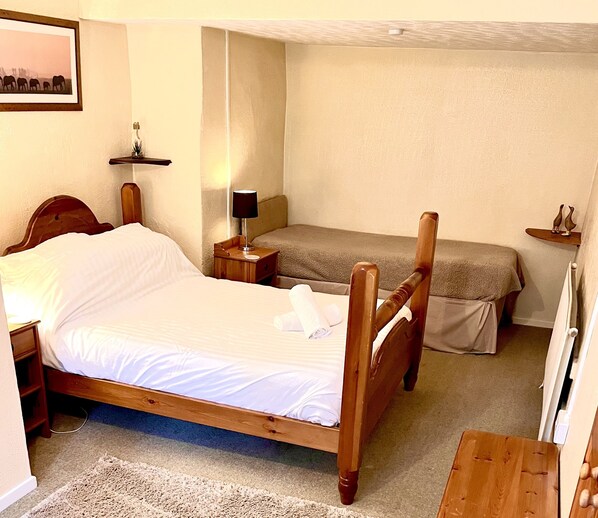 A bedroom with a wooden four-poster bed, a smaller single bed, a bedside table with a lamp, a nightstand, and a small shelf with decorative items.