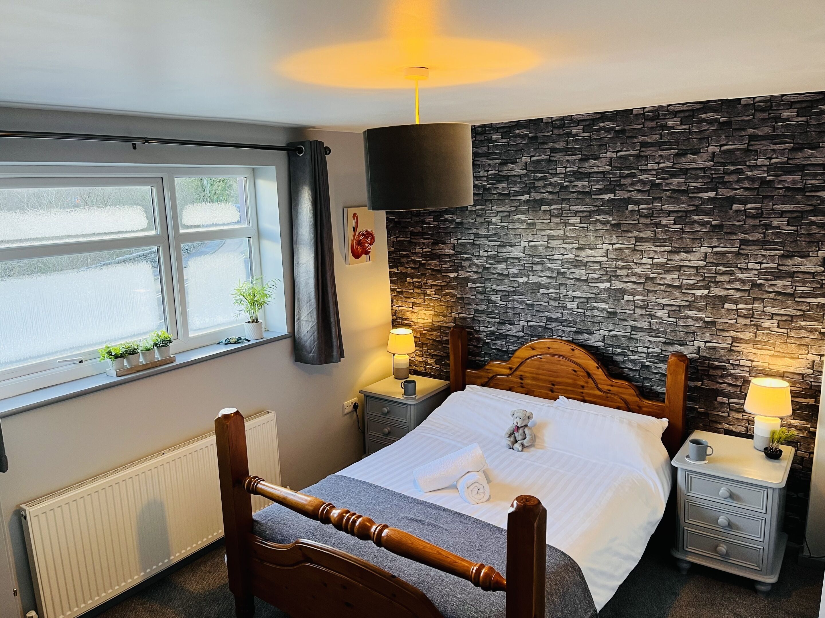 A bedroom with a wooden bed, a stone wall, a window with plants, and a nightstand with a lamp.