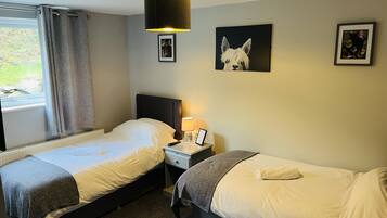 A hotel room with two single beds, a nightstand with a lamp, framed pictures on the wall, and a window with curtains.