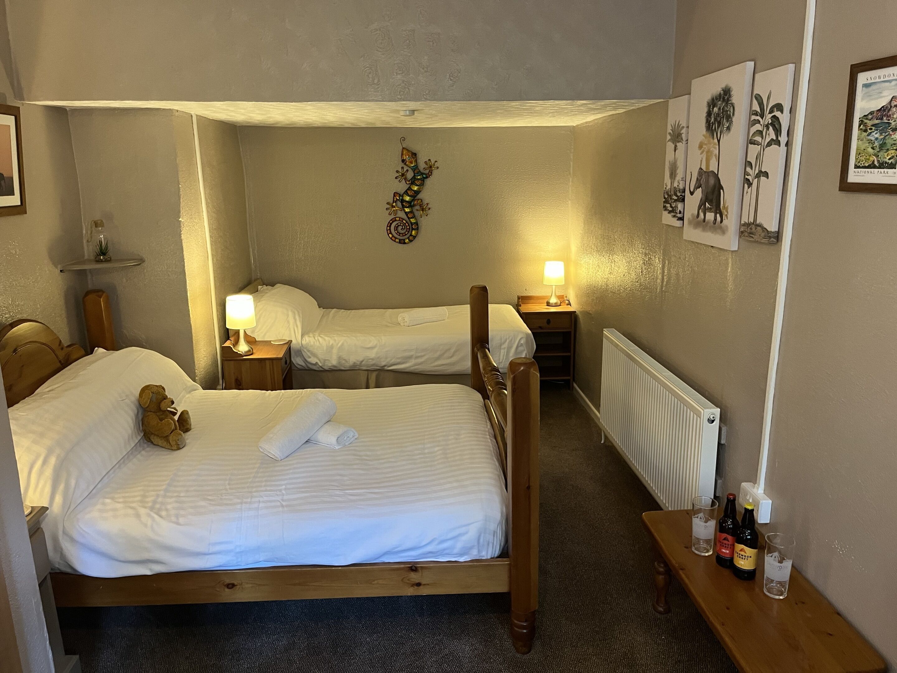 Ett hotellrum med två separata sängar, en sänggavel i trä, ett nattduksbord med en lampa, en hylla med en liten växt, en inramad tavla på väggen och ett element.