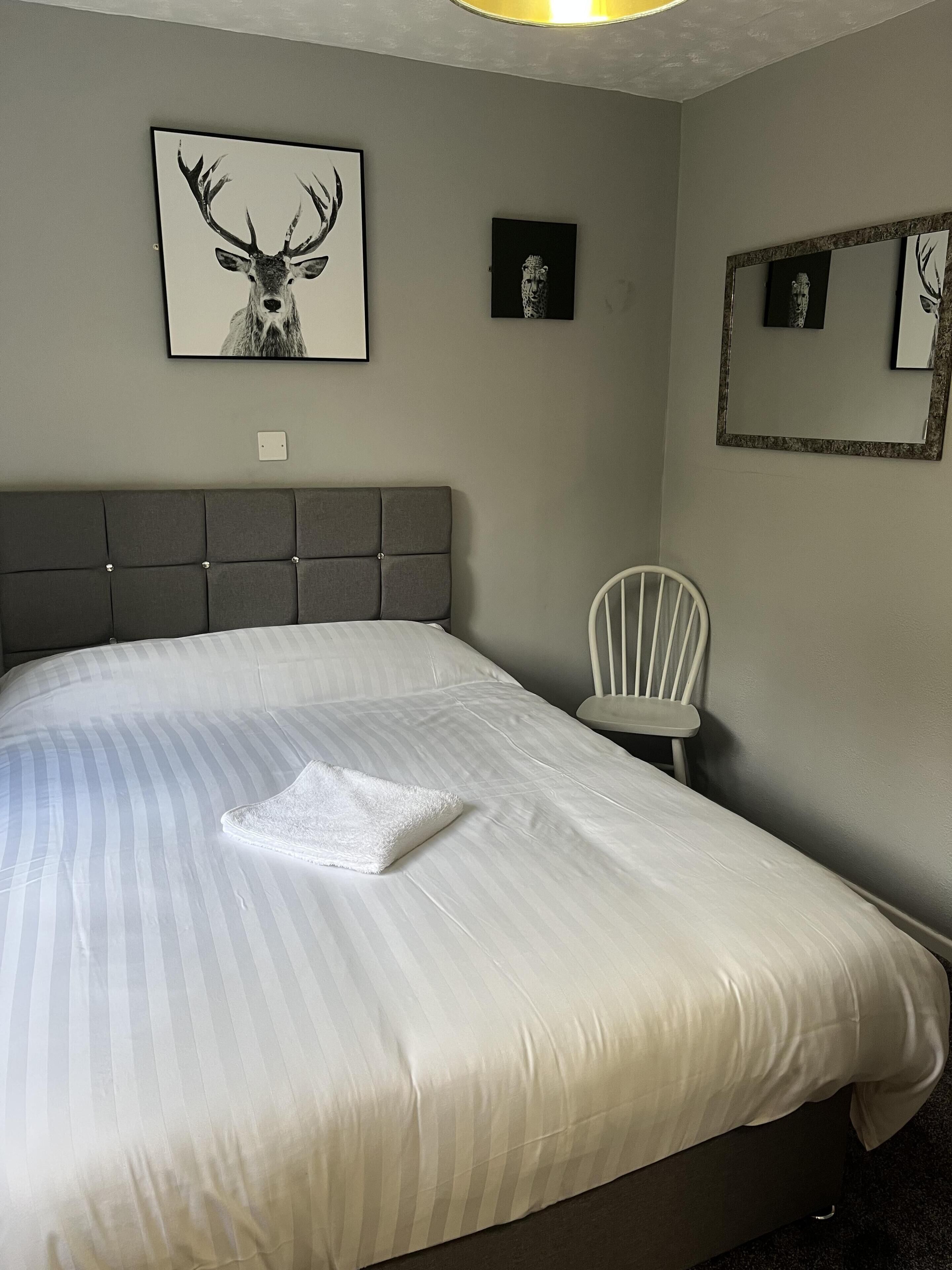 Un lit bien fait, avec une couvre-lit à rayures et une serviette blanche pliée, une tête de lit rembourrée grise, une chaise blanche et des œuvres d’art encadrées accrochées aux murs.